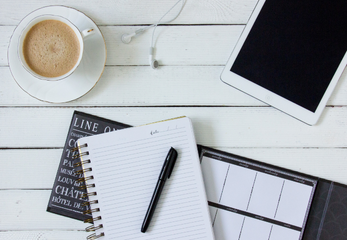 A notebook, calendar, cup of coffee, ear buds, and tablet